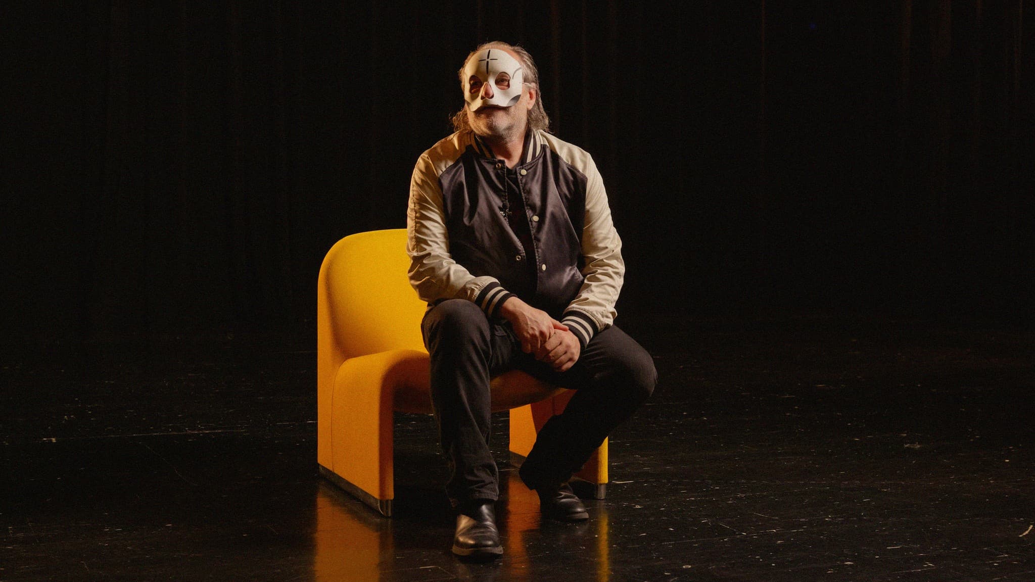 A masked figure sitting on a yellow chair on a dark stage